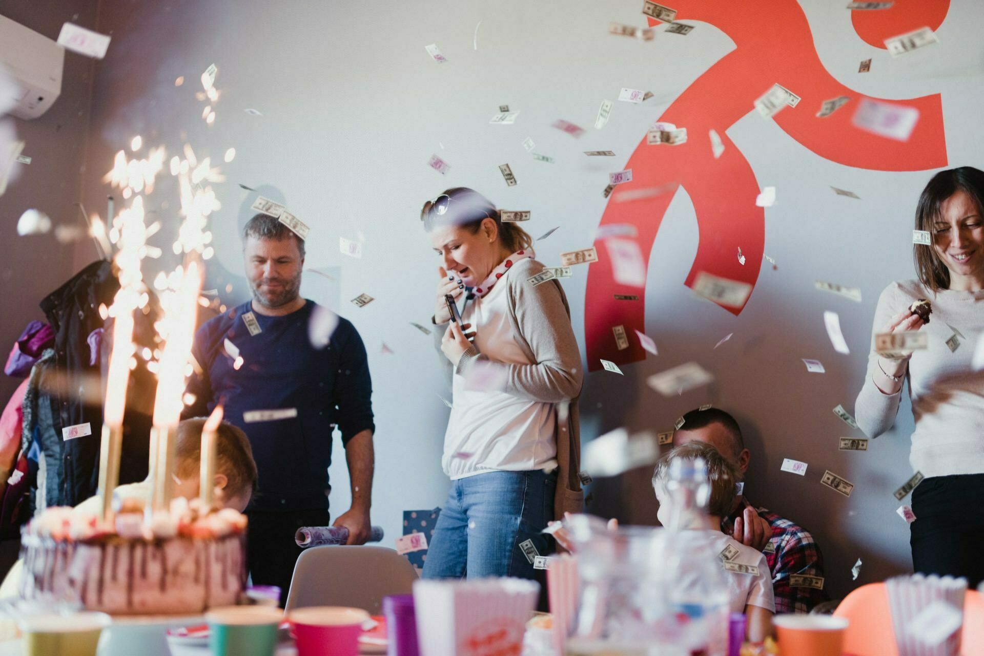 A group of people celebrates with a cake decorated with sparklers, colorful cups and table decorations, and throwing paper money into the air, all expertly captured in a photo report of the events. A large red wall decoration is visible in the background. 