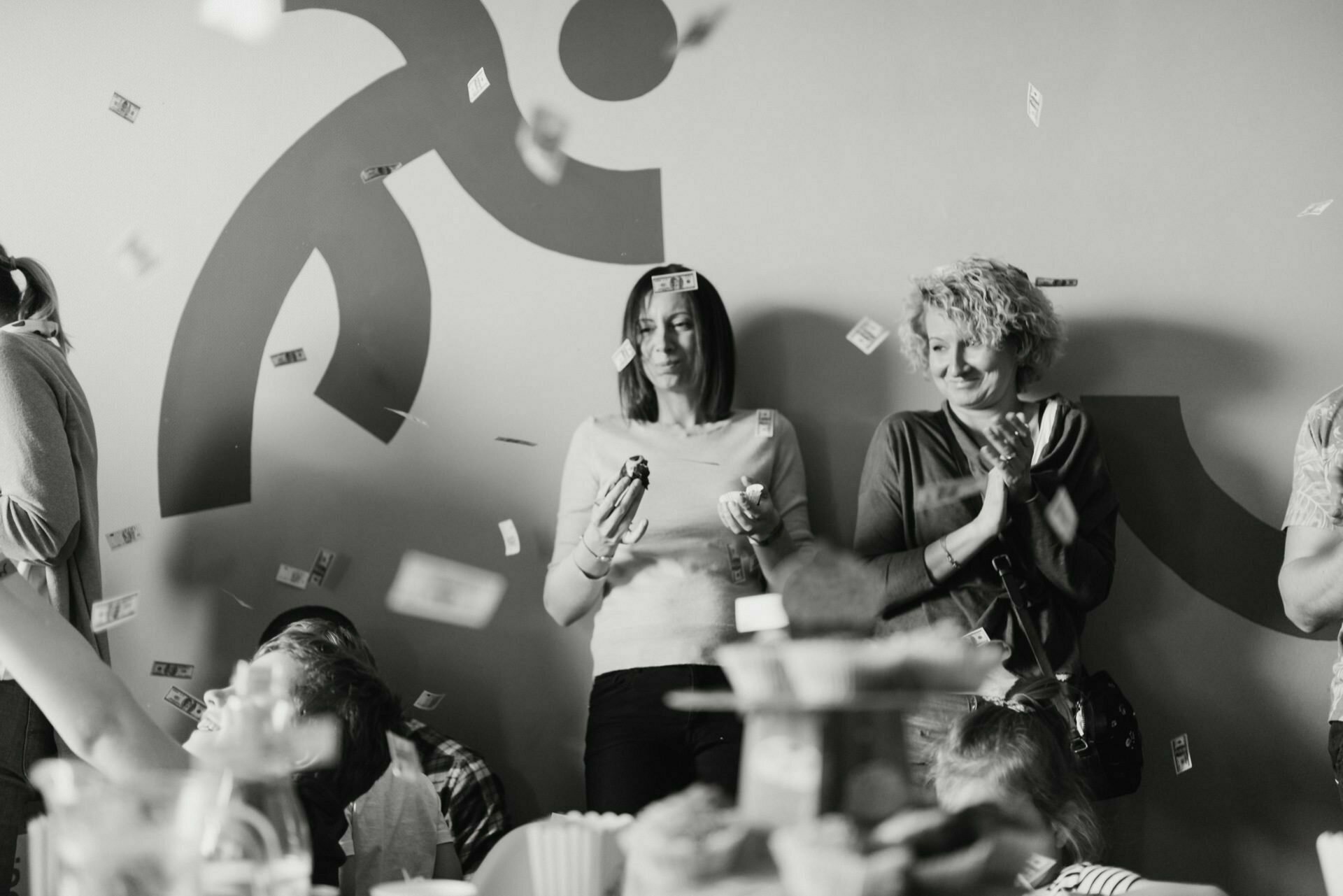 Black and white photo of two women and several children at a party. The women are smiling and clapping as confetti falls around them. An ornate mural is partially visible on the wall behind them, and a table with food and drinks is in the foreground - perfect for a photo recap of the events.  