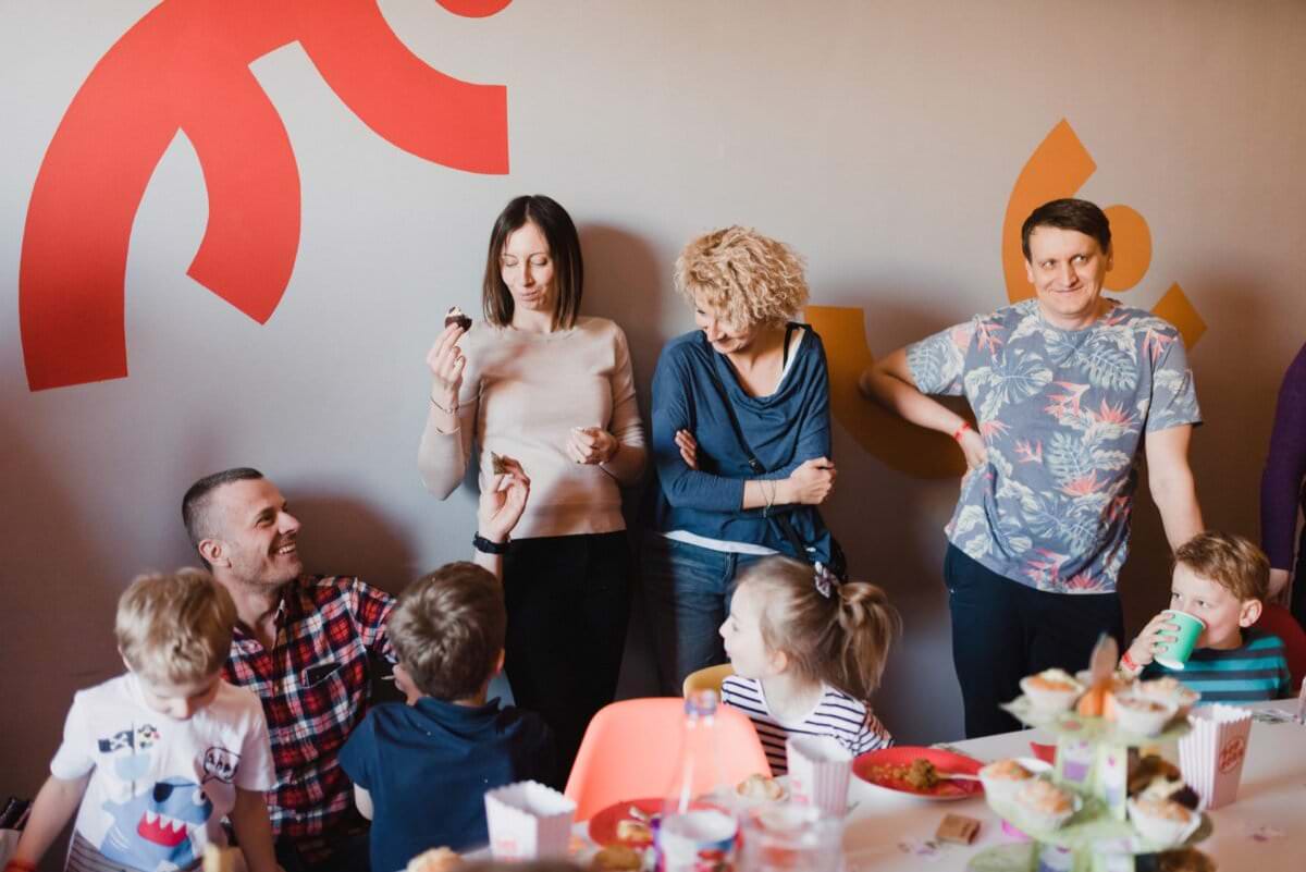 Grupa dorosłych i dzieci gromadzi się wokół stołu z jedzeniem i przekąskami, utrwalając momenty idealne na fotografię eventową. Dwoje dorosłych stoi pod ścianą, jeden trzyma babeczkę. Dwie osoby dorosłe siedzą z dziećmi jedzącymi przy stole. Tętniąca życiem scena obejmuje białą ścianę ozdobioną kolorowymi wzorami, idealną na fotorelację z wydarzeń.