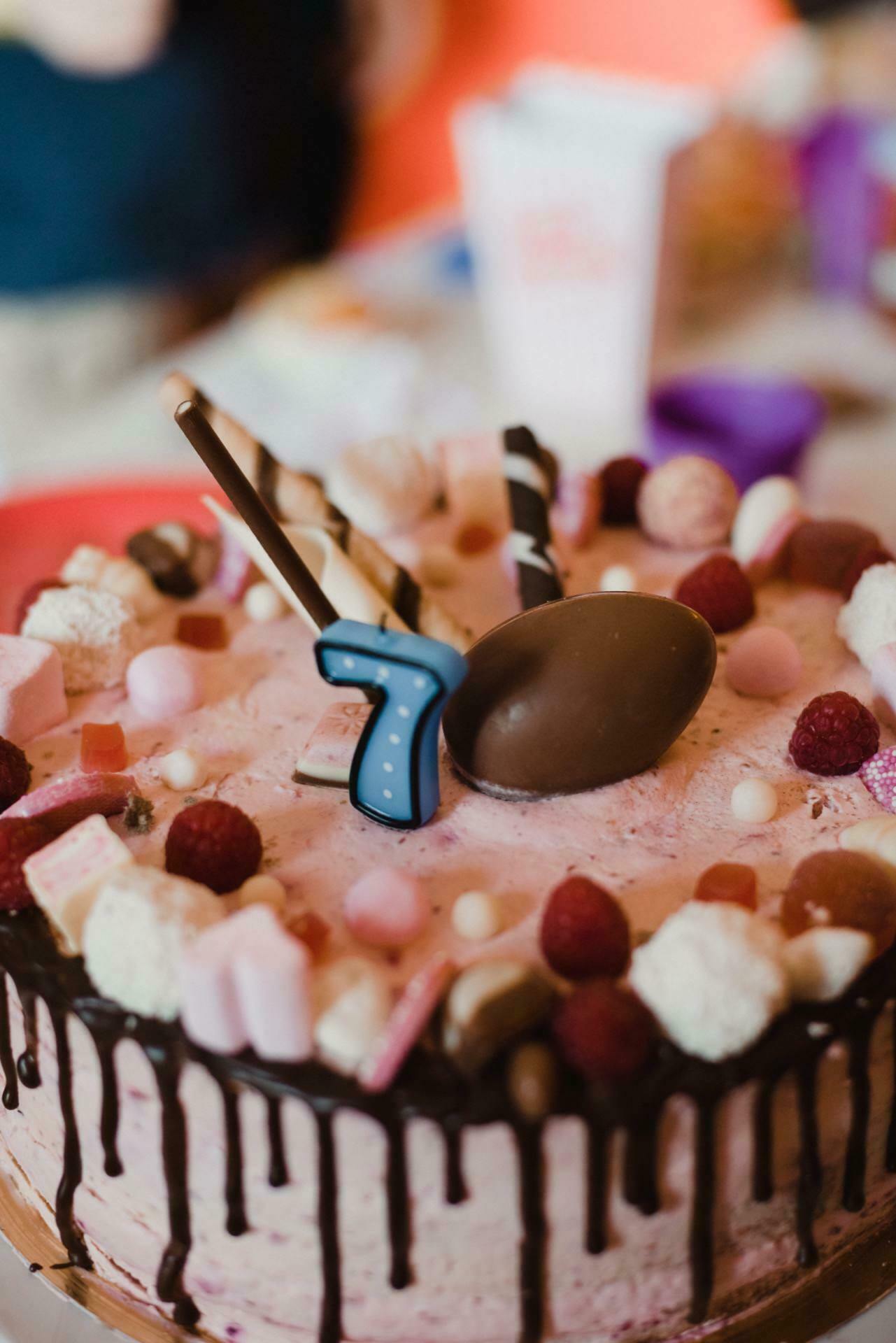 A decorated birthday cake with pink icing, edges dripping with chocolate and various toppings such as marshmallows, raspberries and candies. A chocolate egg and a large blue candle with the number seven were placed on top. This event photo shows a blurry background with party elements.  