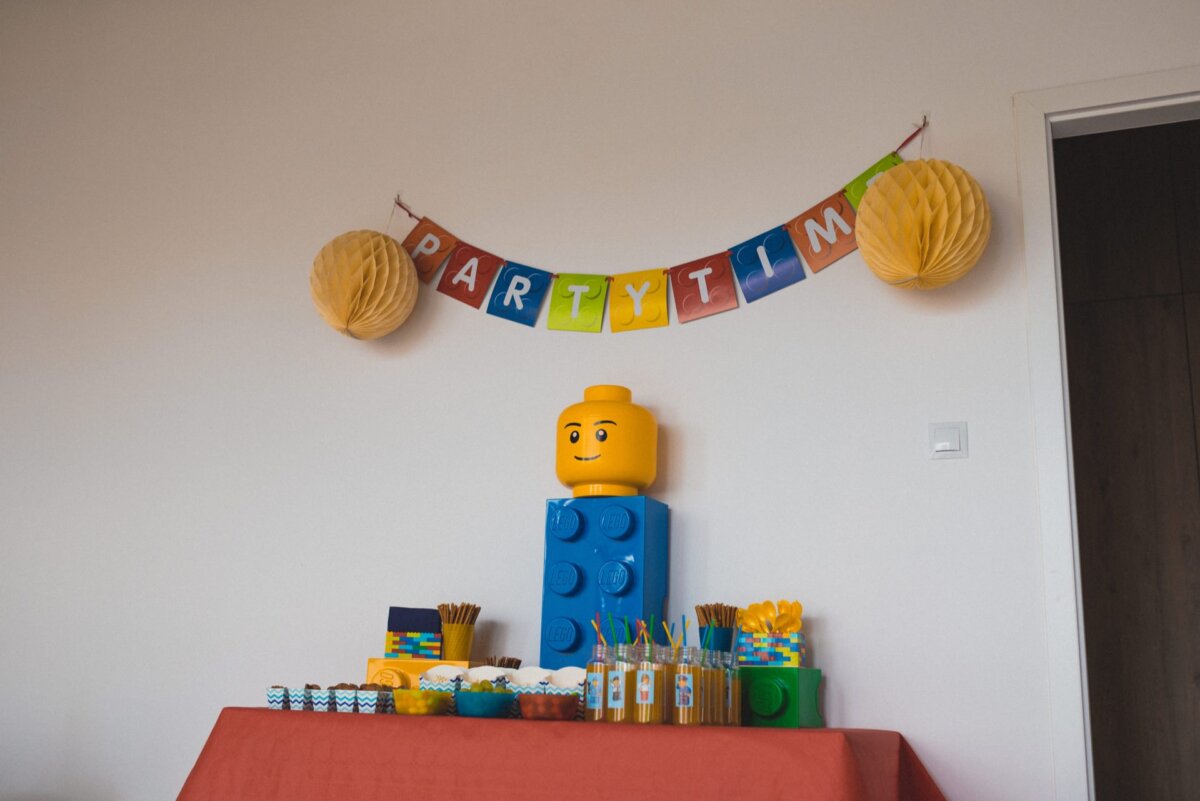 The Lego-themed birthday party set includes a banner spelling out "PARTY TIME" in colorful capital letters. A large yellow Lego head sits atop a blue Lego block on a table adorned with a red tablecloth, surrounded by a variety of snacks and decorations - all captured in exquisite detail in event photography for an unforgettable photo event. 