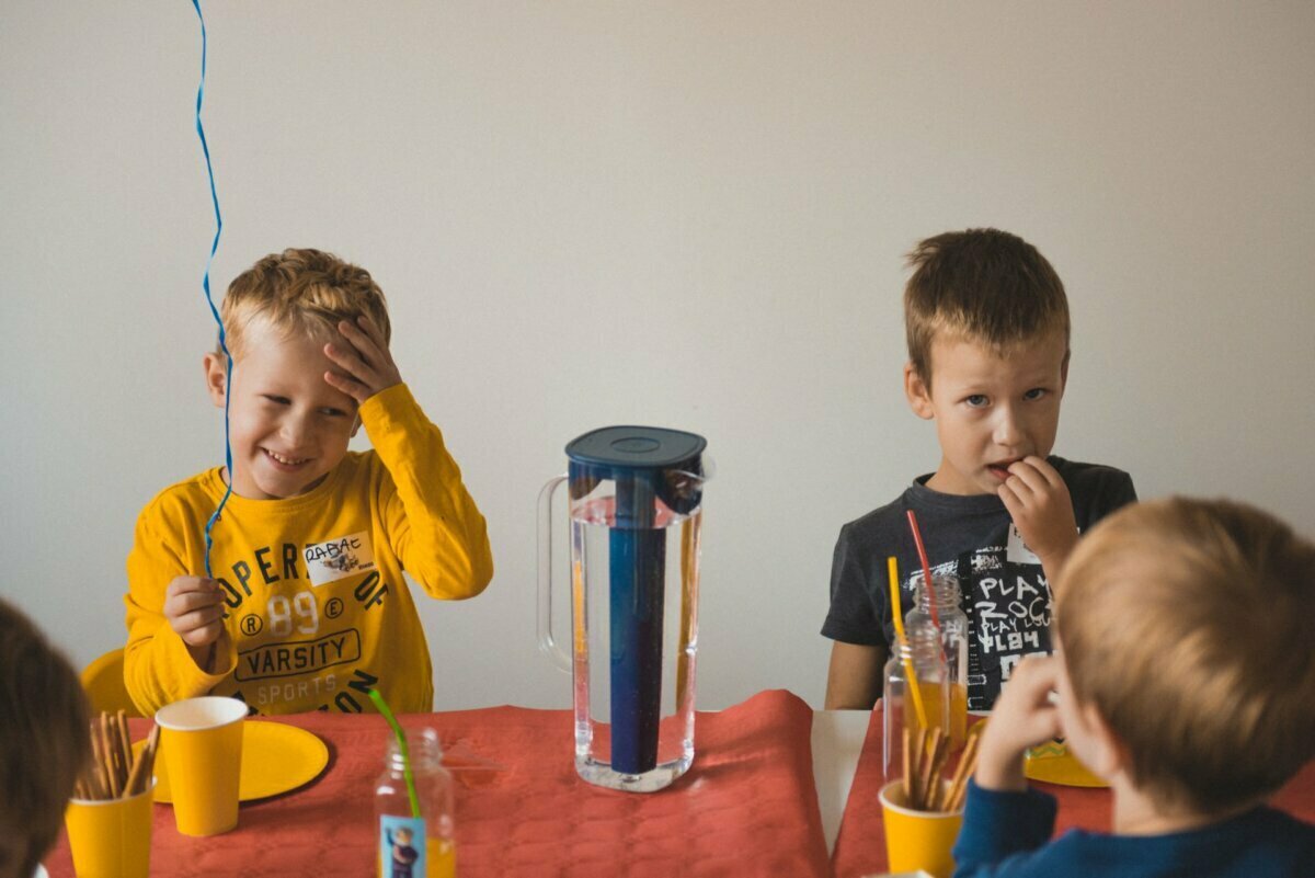 Two children sit at a table decorated for a party. The child on the left, wearing a yellow T-shirt, is smiling and holding a balloon. The child on the right, dressed in a dark T-shirt, looks thoughtful while eating. On the table are party supplies, snacks and drinks - capturing the perfect event photography moment.   