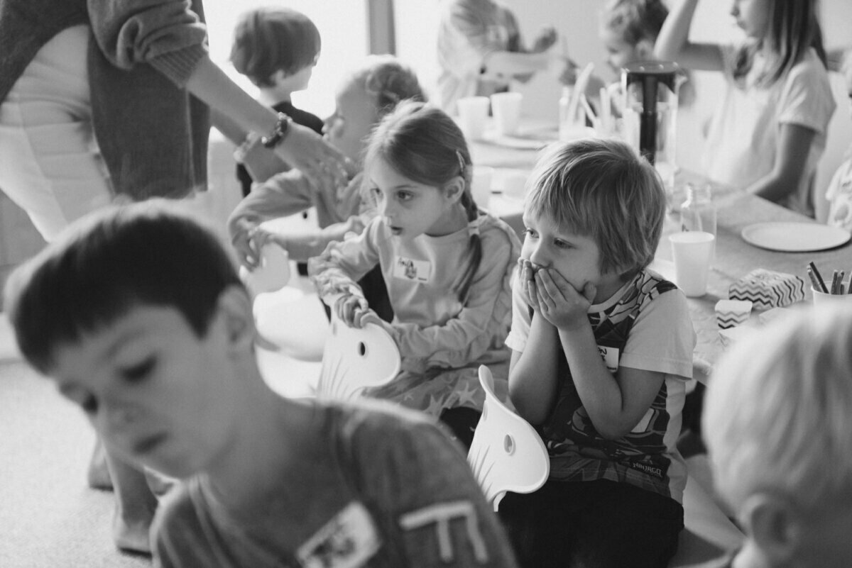 Black and white image showing a group of young children sitting around tables and engaged in some activity. In the foreground, a boy with his hands covering his mouth looks attentive while another child listens. In the background are other children and an adult, perfectly capturing this event photography.  