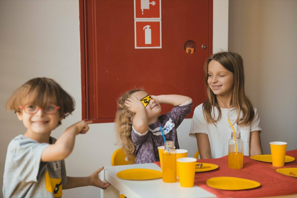 Three children sit at a table set with yellow cups and plates in a room with a red wall. One child points, the other covers his eyes with a napkin. The third child smiles and watches the others, with a bottle and straw placed on the table, capturing the perfect moment for any event photography.  