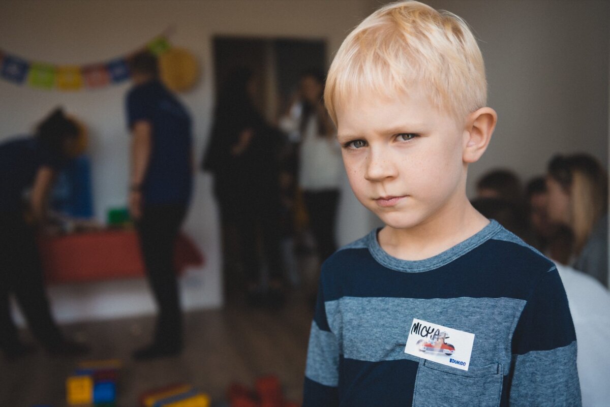 A young boy with blond hair stands in a room, wearing a blue striped shirt and a badge sticker. The background is slightly blurred, capturing the essence of event photography, with people engaged in activities and colorful decorations on the wall. The boy is looking directly into the camera with a serious expression on his face.  