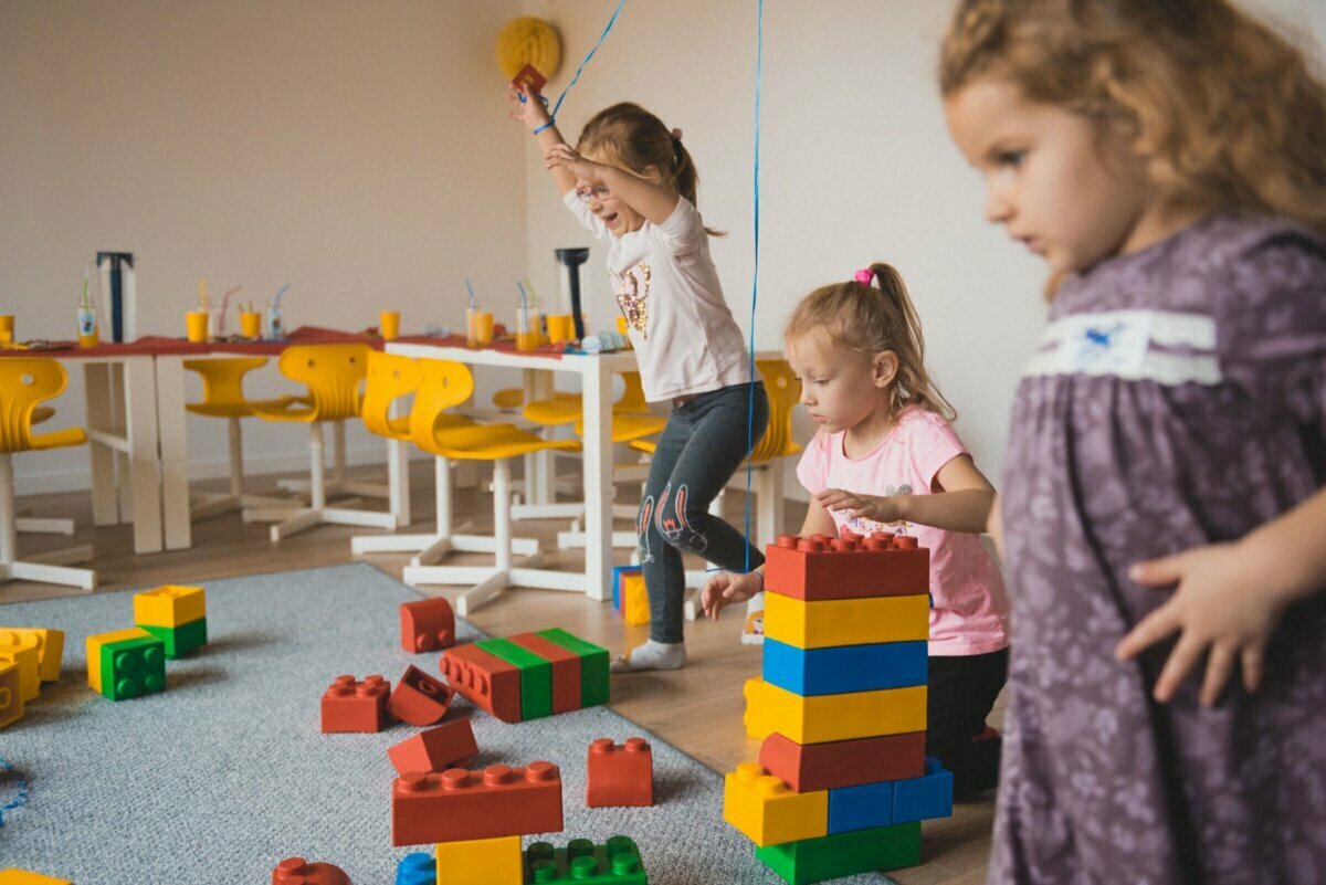 Three young girls play with large colorful blocks on a carpeted floor in a brightly lit room. In the background are yellow chairs and tables with snacks and drinks. One girl is jumping with excitement, the other two are focused on building - perfect for photo coverage of the events.  