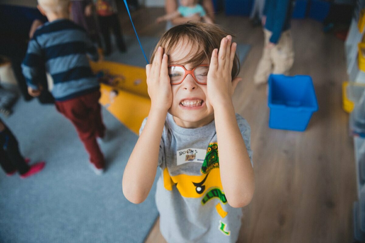 A child with brown hair, wearing glasses and a gray T-shirt, playfully covers his eyes while smiling. In the background, other children are engaged in activities at the colorful indoor playground, which is perfectly captured in this vibrant photo report of the events. 