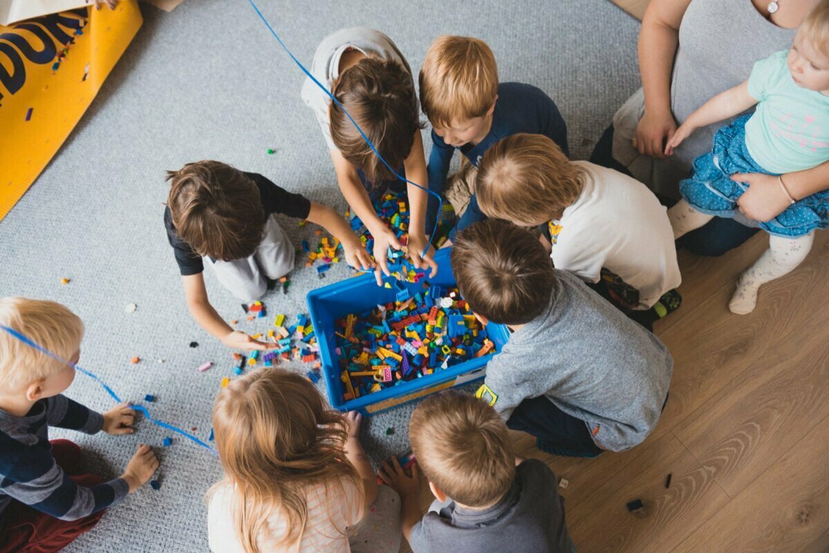 A group of children sit on the floor and surround a blue basket filled with colorful blocks. They enthusiastically reach into the basket, and some are already holding the blocks. The scene seems playful and cooperative, perfect for a photo essay of the events, capturing their joyful interactions.  