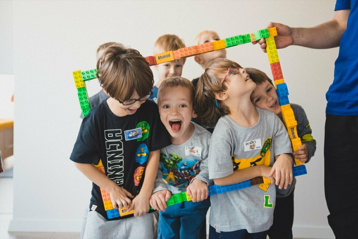 A group of smiling children cluster together, holding a colorful Lego frame around them. Some are wearing glasses, and all seem happy and engaged. An adult hand on the edge of the frame helps hold it up. This event photography, taken in Warsaw, has a bright, minimalist background.   