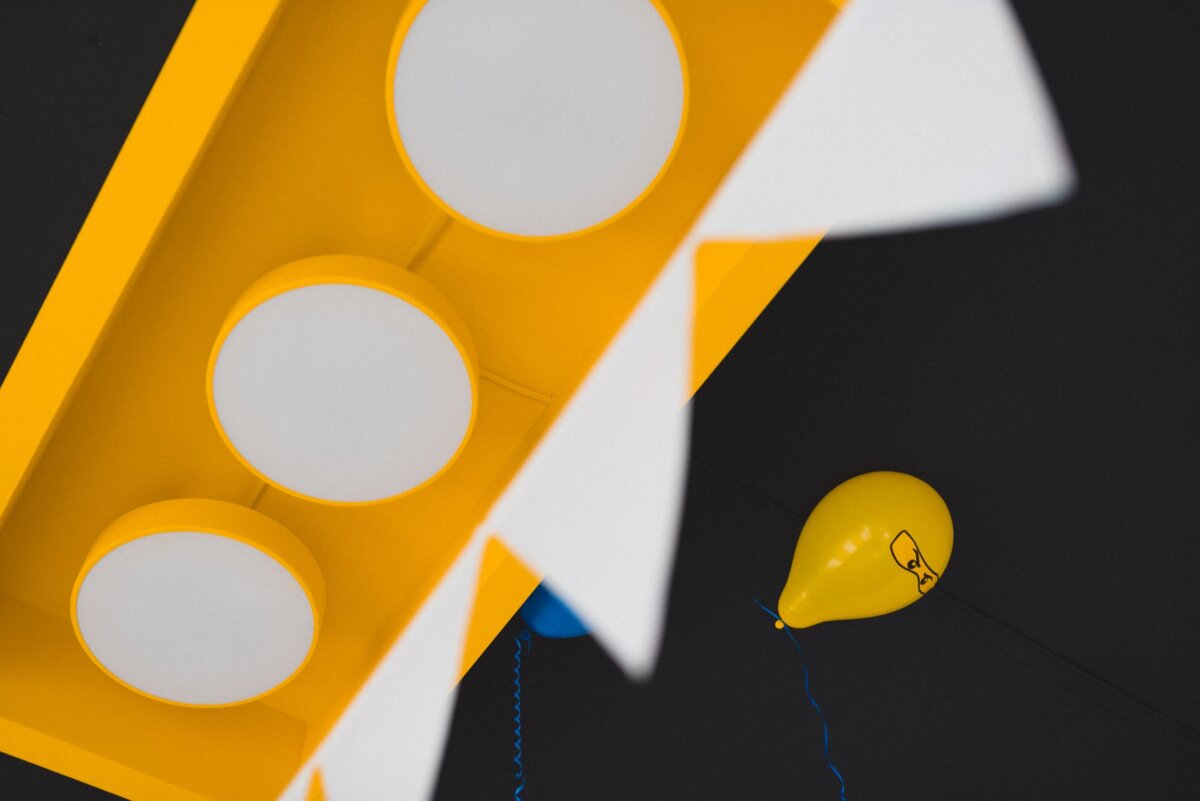 A yellow ceiling fixture with three round white bulbs can be seen from below. White triangular pennants hang in the foreground, and under the fixture a yellow balloon with a cartoon face floats on a dark background, capturing the essence of event photography. 