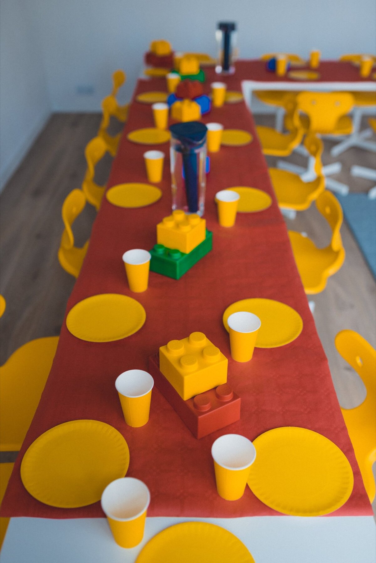 A long table set for a children's party includes a red tablecloth, yellow paper plates and yellow cups. Decorated with large Legos and surrounded by yellow chairs, the scene is perfect for capturing memorable moments through event photography. 