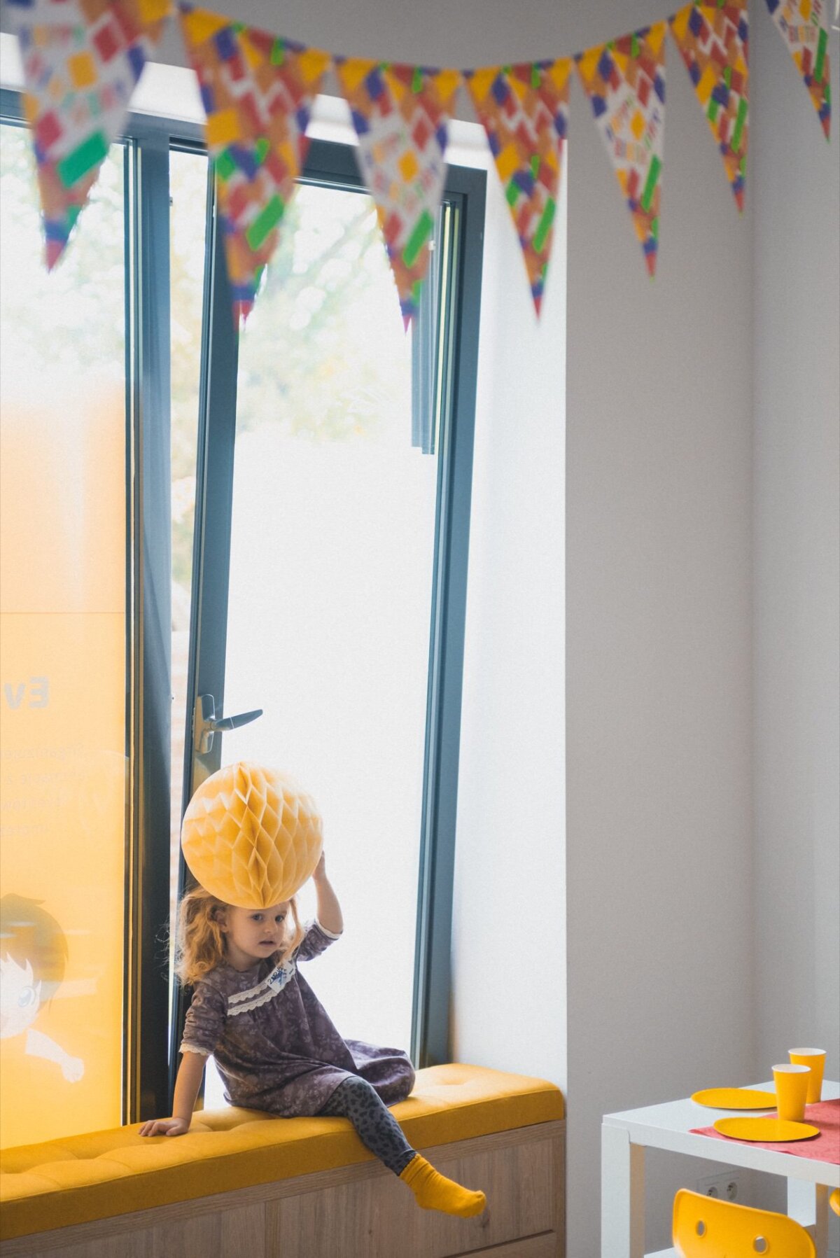 A young child sits on a yellow padded bench by the window, holding a yellow paper honeycomb decoration on his head. The room captured in this delightful photo-reportage of events is decorated with a colorful triangular flag hanging from the ceiling. A table with yellow cups and plates is partially visible in the foreground.  