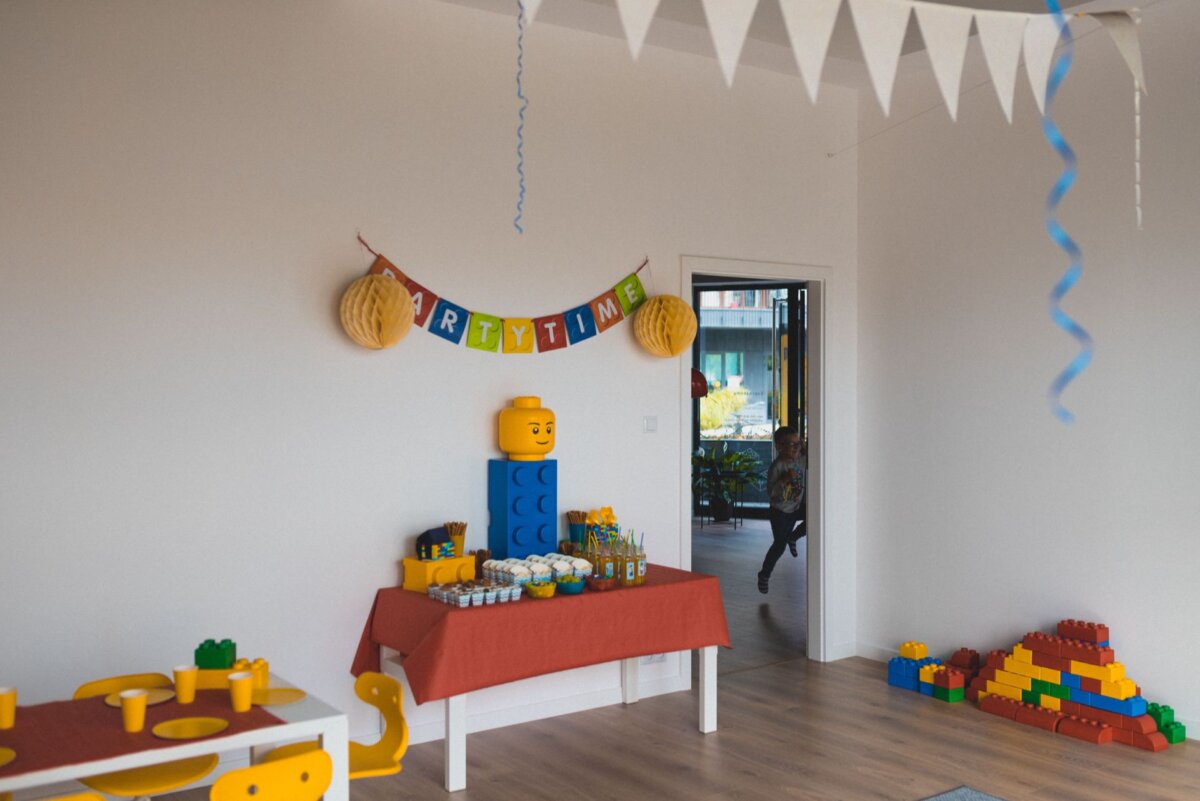 The room decorated for the children's party includes a "Time to Party" banner, colorful streamers and LEGO-themed decorations. A table with a red tablecloth holds snacks and drinks, while a boy runs around in the background. The floor is strewn with LEGO bricks and toys, perfectly capturing the mood of the photo event.  