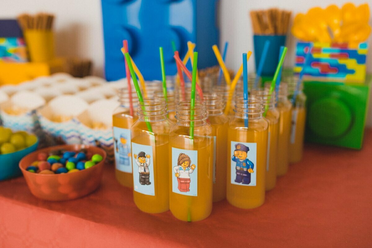 On the table are arranged bottles of orange juice with colorful straws and stickers with LEGO characters. Bowls with colorful candies stand next to them. The table is decorated with LEGO-themed items and snacks, perfect for vibrant event photo coverage by any event photographer Warsaw.  