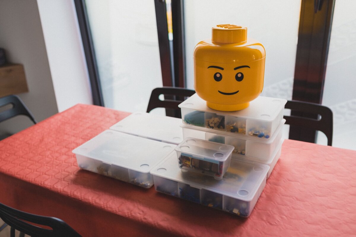 A stack of clear plastic storage containers filled with various LEGO pieces is set on a table covered with a red tablecloth. At the top of the stack is a large yellow LEGO head storage container with a smiling face, capturing the playful essence perfect for any "photo event." 