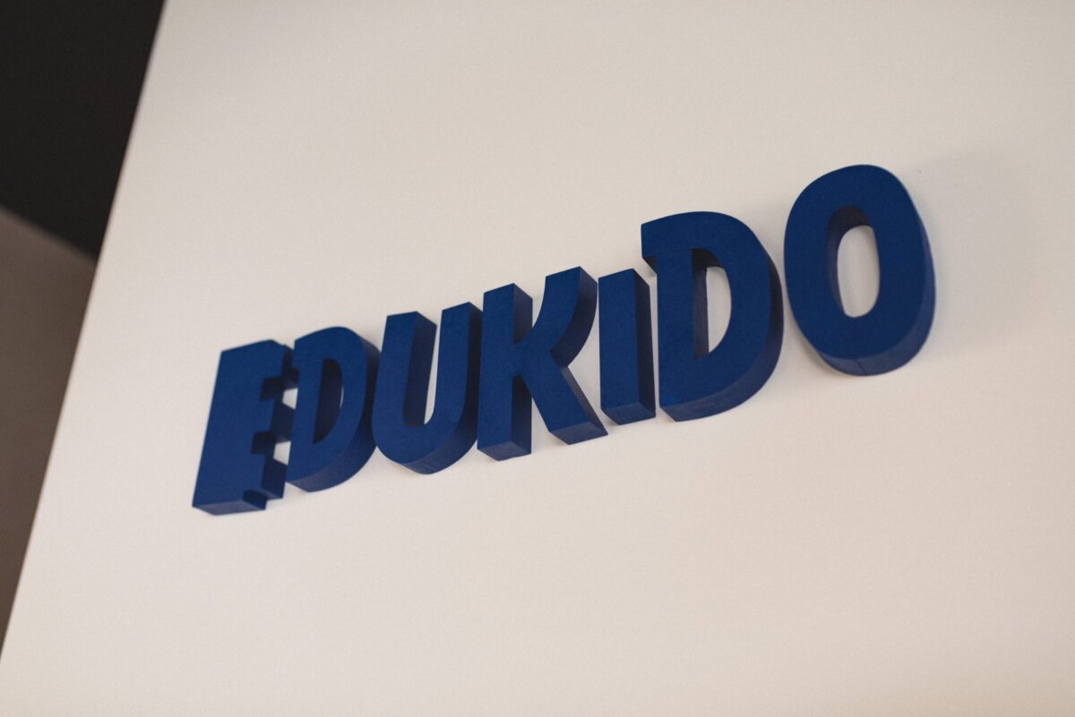 Blue, three-dimensional letters spelling "EDUKIDO" are mounted on a plain wall in broken white. The design has a clean, modern look, perfect for framing event photography. 