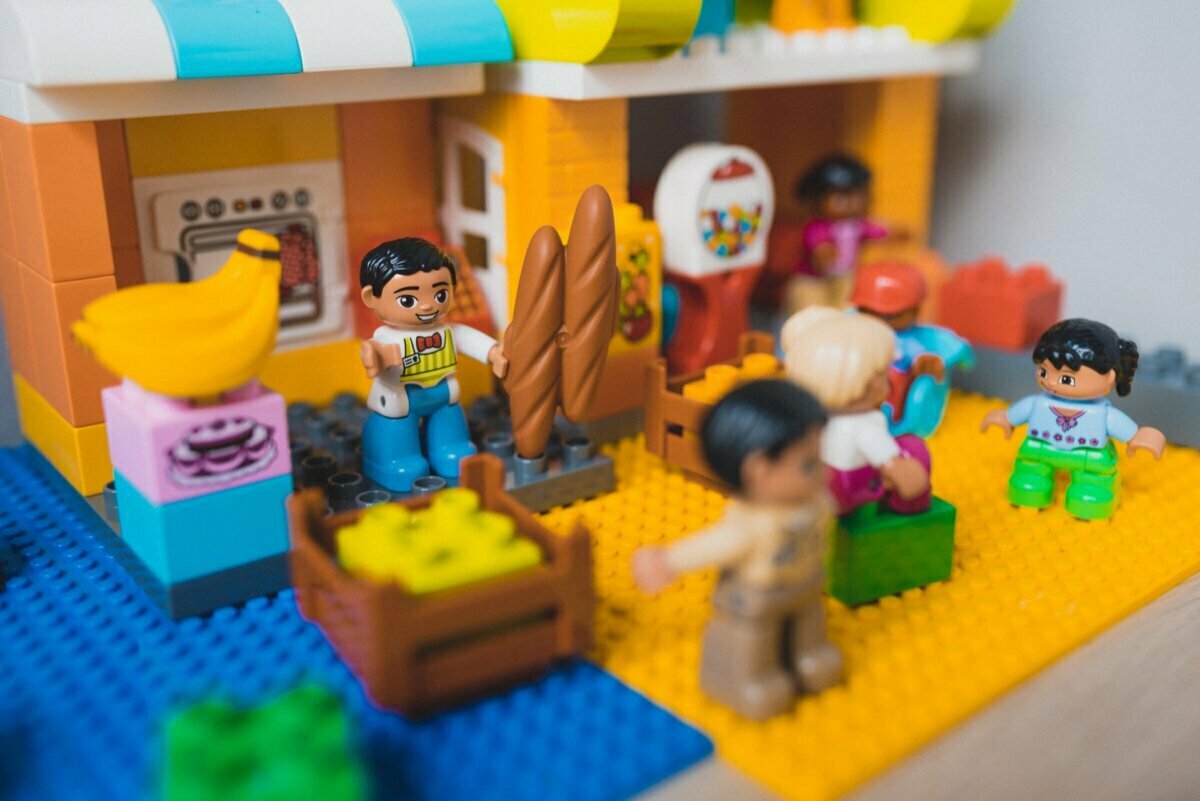 The colorful scene from the LEGO set captures the vibrant moment of the event photographer warsaw: one figure holds baguettes, another cradles a baby, and others perform various activities. A miniature building, a bunch of bananas and a variety of small props complete this delightful event photographer. 