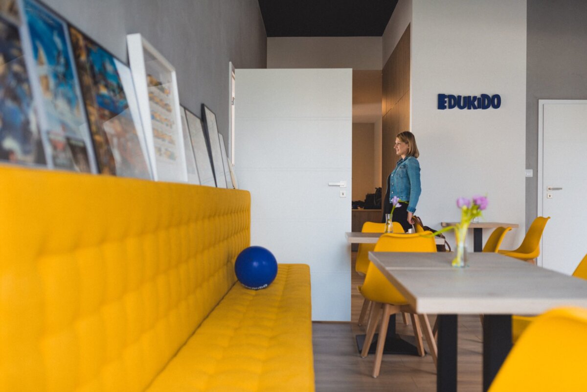 The person is standing in the open doorway of a modern room with yellow padded benches and chairs, wooden tables and colorful wall decorations. On the bench is a blue ball and a small vase of flowers. A sign in the room reads "EDUKIDO." This scene could be included in an event photo story by event photographer Warsaw.   