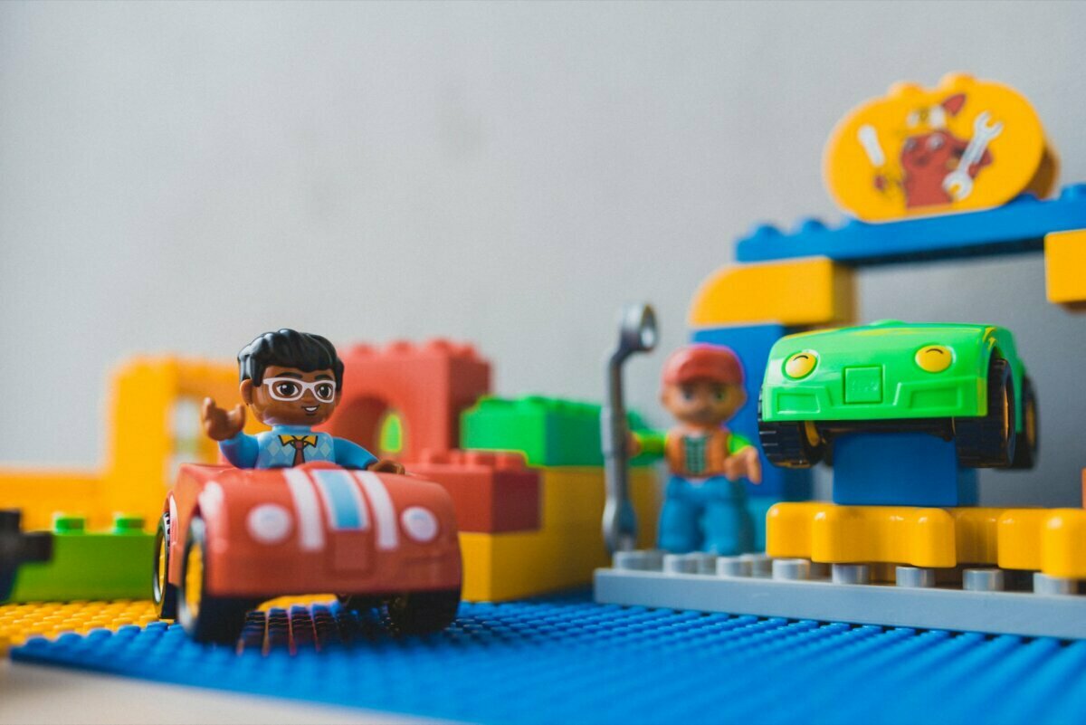 Colorful LEGO scene with two minifigures: one driving a small red car, and the other holding a key near a raised green car. In the background are various LEGO bricks and a structure with a crab emblem at the top, perfect for capturing in the next photo story of the events. 