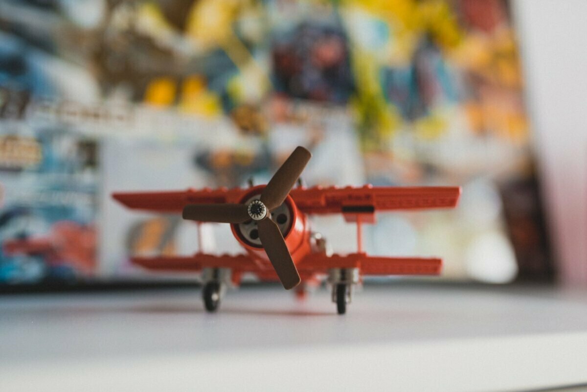 A red toy biplane with brown propellers is displayed on a white surface. The background is blurred and includes colorful illustrations or posters, creating a vivid backdrop for the small model airplane, much like an event photograph showing the essence of the event. 