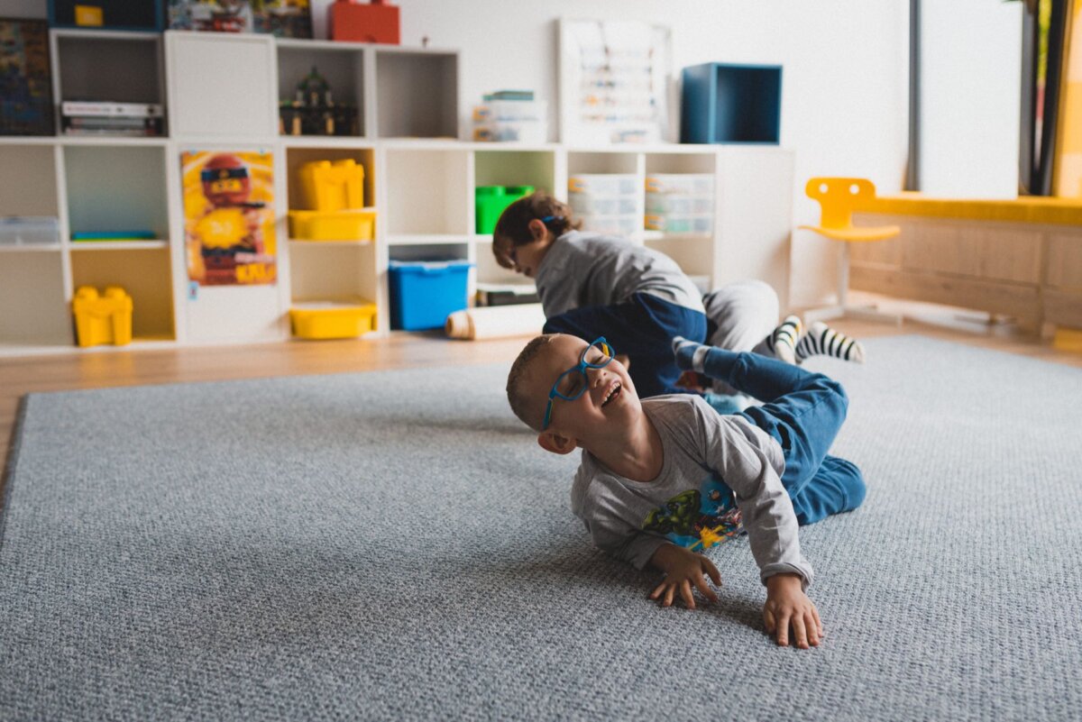 Two children roll over on a gray carpet in a well-lit room for fun. One child wearing glasses and a gray shirt is laughing in the foreground. There are various toys and containers on shelves in the background, and a yellow chair stands next to the window - a perfect scene for event photography.  