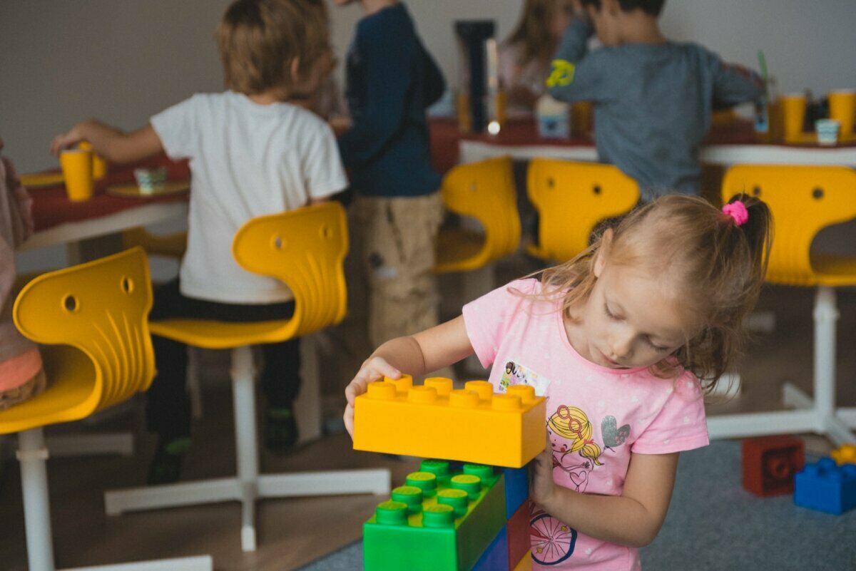 A young girl in a pink shirt is playing with large colorful blocks in the foreground. Other children are sitting at a table with yellow chairs in the background doing various activities. This charming scene, captured by event photographer warsaw, highlights the happy atmosphere in the classroom.  