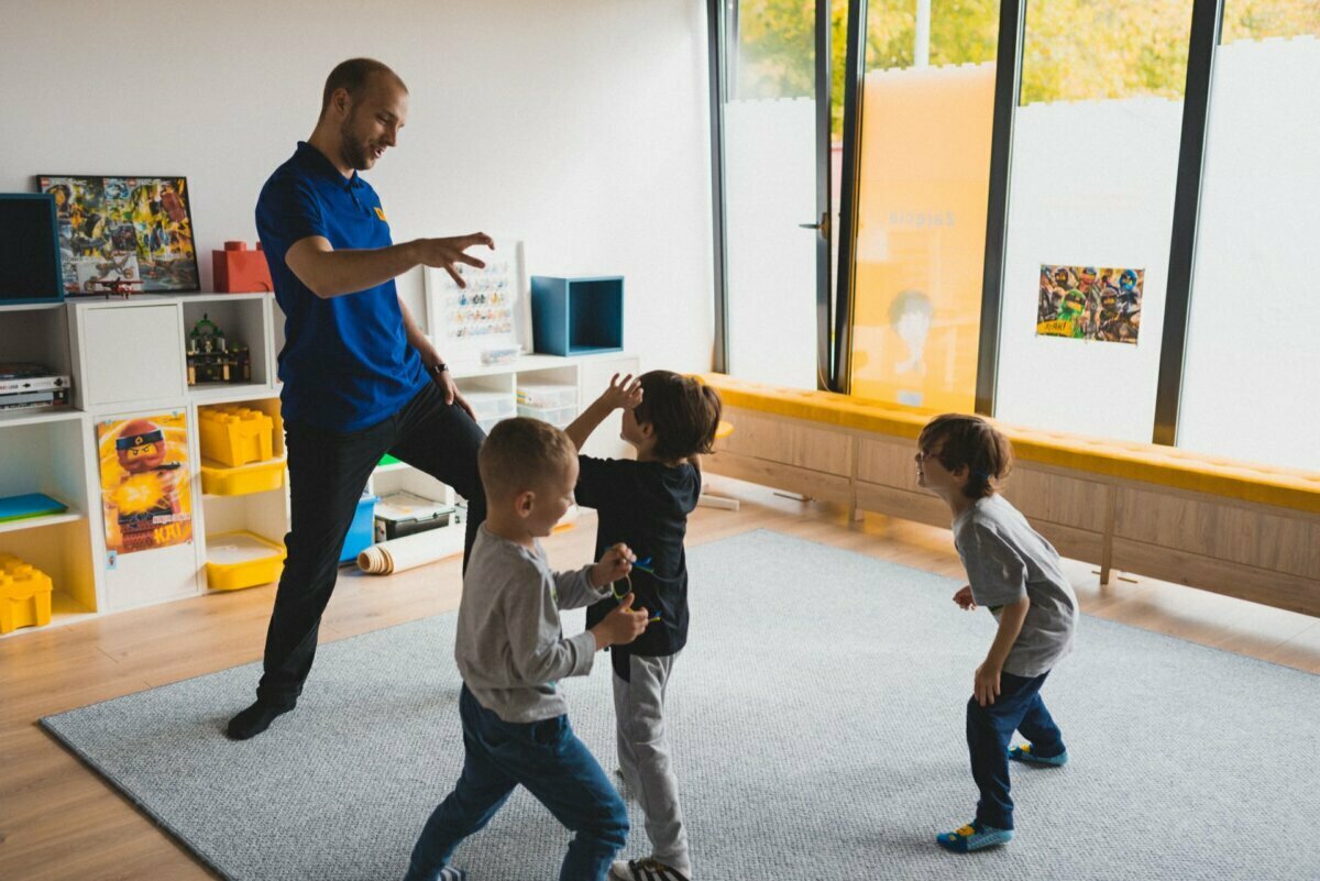 A man in a blue shirt is playing with three young children in a brightly lit room filled with various toys. The children are engaged and you can see that they are having fun. The room has large windows and colorful decorations on the walls, creating the perfect setting for event photography.  