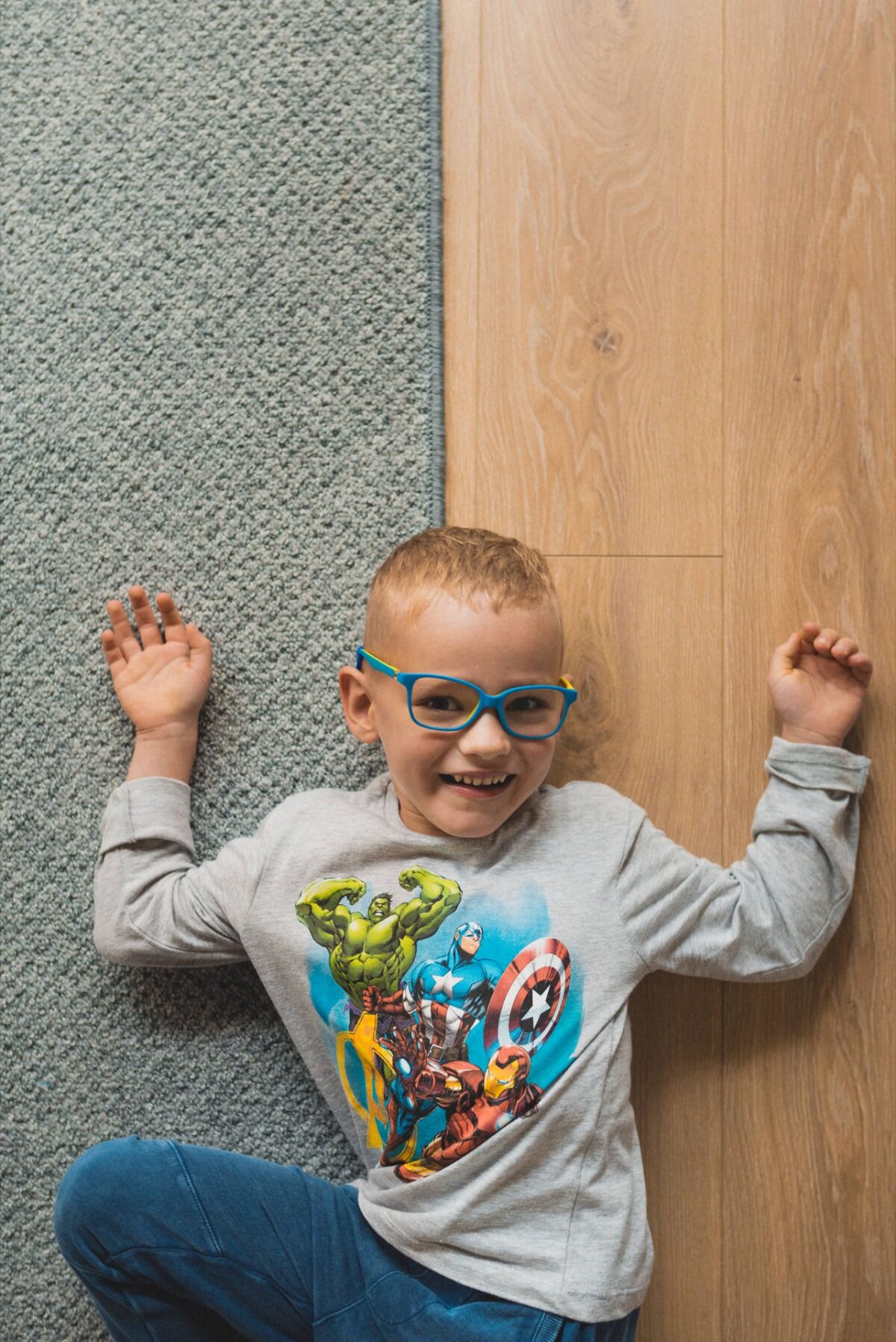 A young child with short hair and blue-rimmed glasses lies on the floor during an event photo, with one hand on the gray carpet and the other on the wooden floor. The child is smiling and wearing a gray T-shirt with superhero characters, capturing the joy in the event photo. 