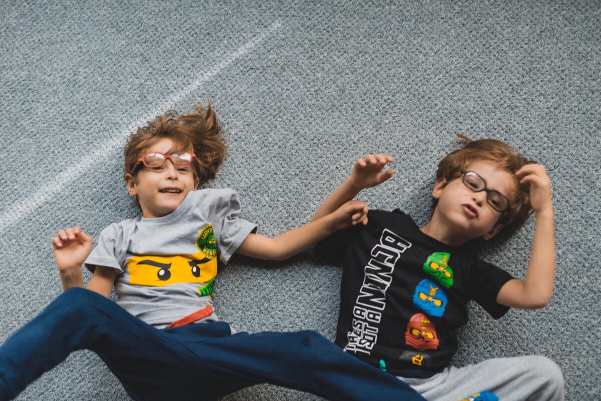 Two children with glasses are lying on a carpeted floor and playfully posing. The child on the left is wearing a gray T-shirt with a graphic depicting a LEGO face, while the child on the right is wearing a black T-shirt with colorful cartoon characters. This joyful moment could easily become part of an event photo shoot.  