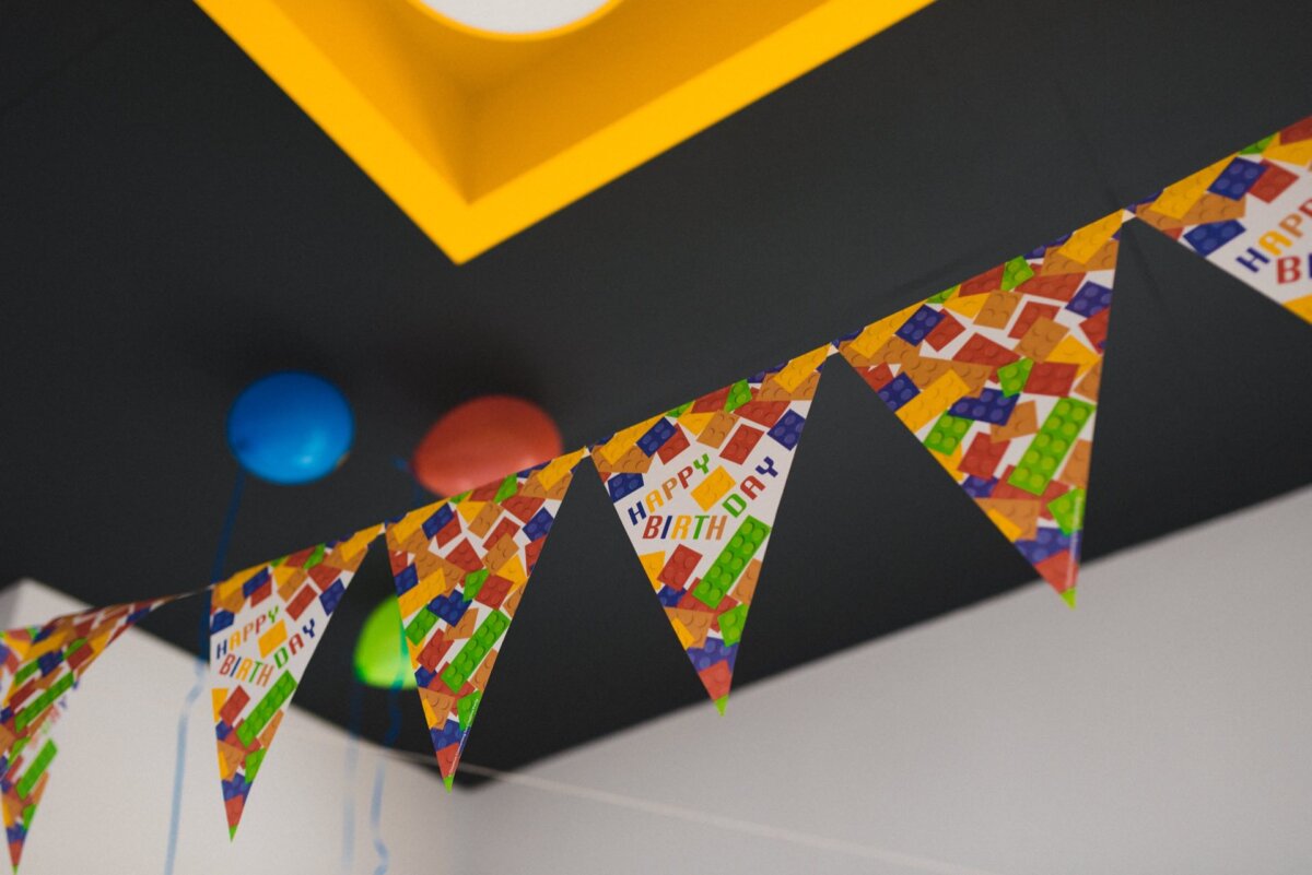 A room decorated for a birthday party with colorful triangle banners with pennants reading "Happy Birthday." Two floating balloons, one blue and one red, float from the ceiling with a yellow light fixture, perfect for event photography to capture every joyous moment. 