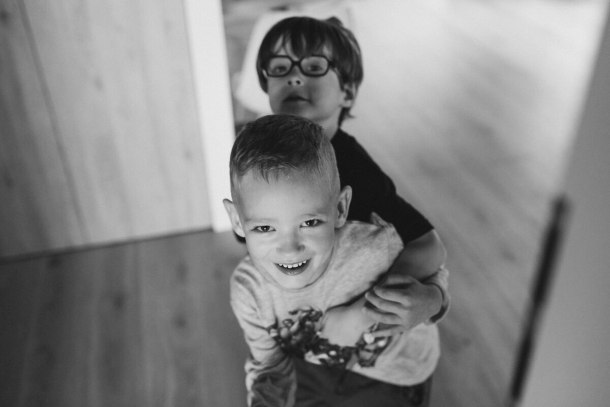 Black and white photo of two young boys in a room. The boy in front, smiling and looking into the camera, is hugged from behind by the other boy with glasses. They are standing on a wooden floor near a doorway with light streaming in from the side, captured by an event photographer warsaw.  