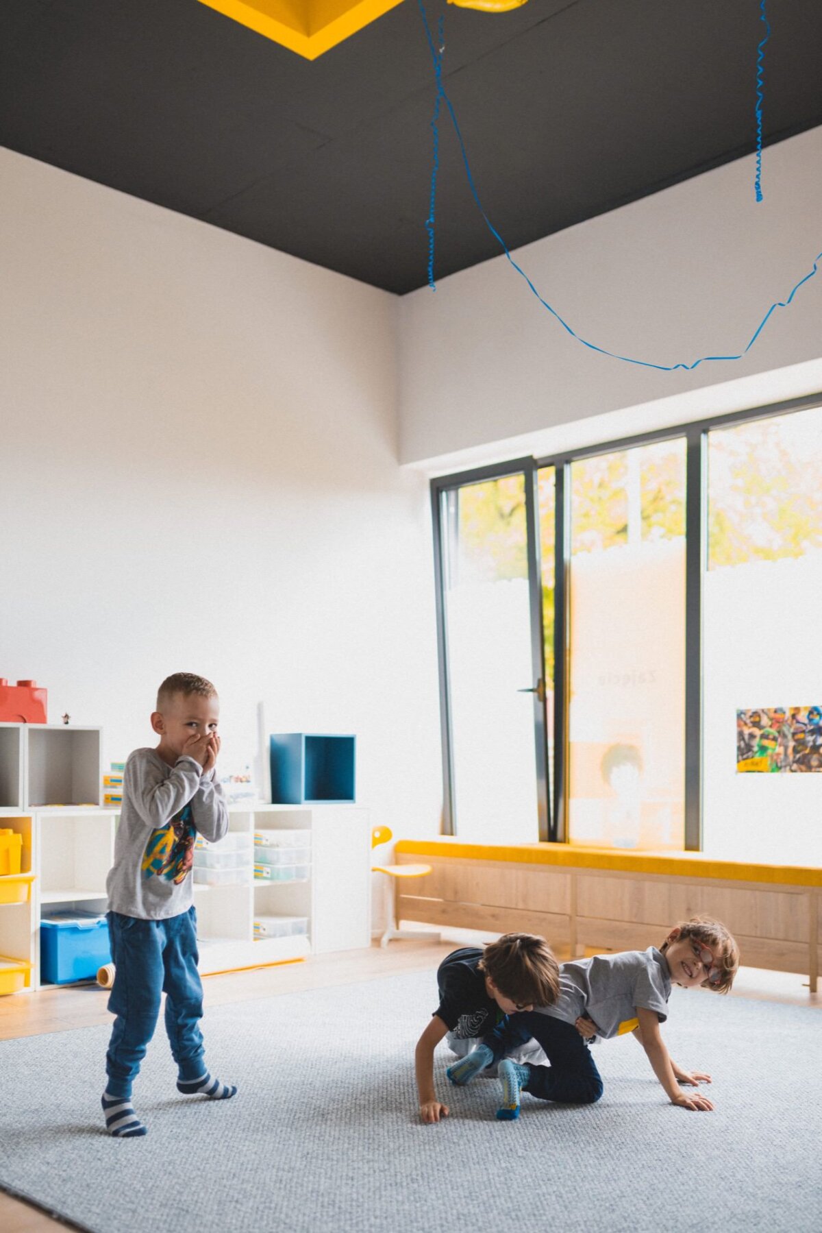 Three children are playing in a bright room with large windows. One child stands alone on the left side, smiling and covering his face with his hands. The other two children lie on the floor and joke around. In the background, shelves of toys can be seen, perfectly capturing the moment of the photo-relation of events.   