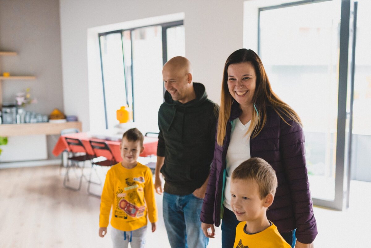 A smiling family of four stands in a bright room with large windows. The parents, a man in a green hooded sweatshirt and a woman in a purple jacket, stand behind their two little boys in yellow T-shirts. A dining table with chairs is visible in the background, typical of event photography.  