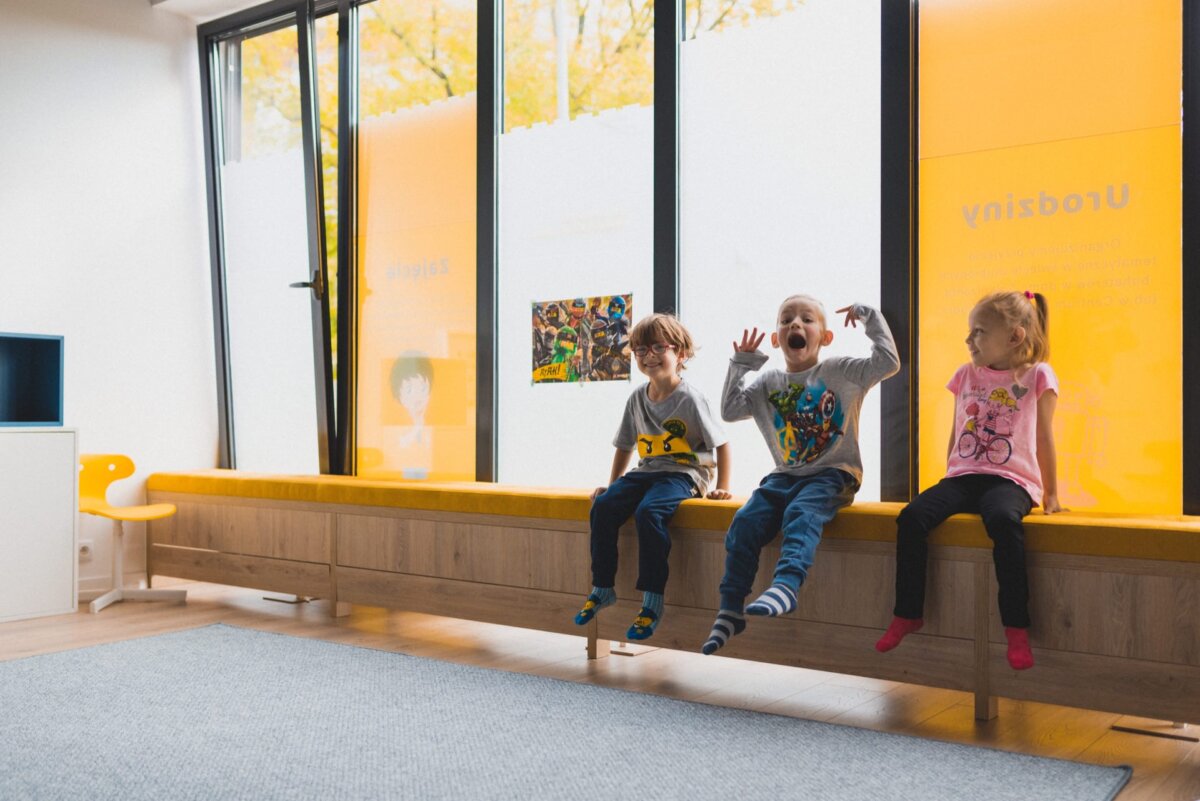 Three children are sitting on a long yellow bench in a bright room with large windows. Captured by the event photographer Warsaw, they appear to be in a playful mood; one child has his hands raised and his mouth open, as if making a funny face. The room has a minimalist design, and the windows let in lots of natural light.  