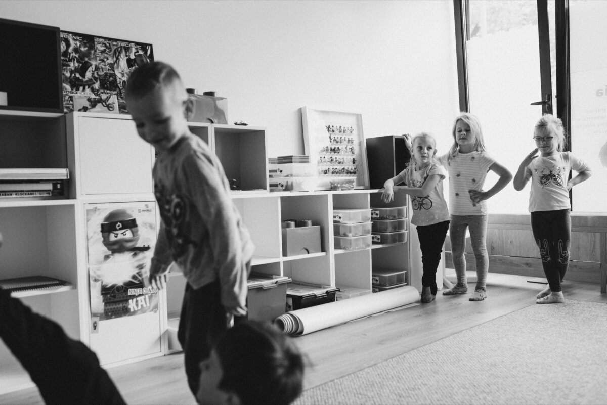 Black and white image of several children in a classroom. Four children stand in a row against a wall with shelves of books and toys, with one child moving in the foreground. Posters and boxes hang on the shelves, and a door is partially visible in the background - captured by event photography.  