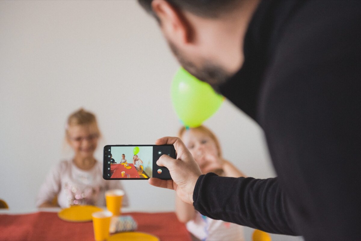 A man dressed in a black sweater takes a cell phone photo of two smiling women sitting at a table. The table is set with yellow cups, plates and a red tablecloth. One of the women is holding a green balloon, capturing the essence of the vibrant photo of the events.  