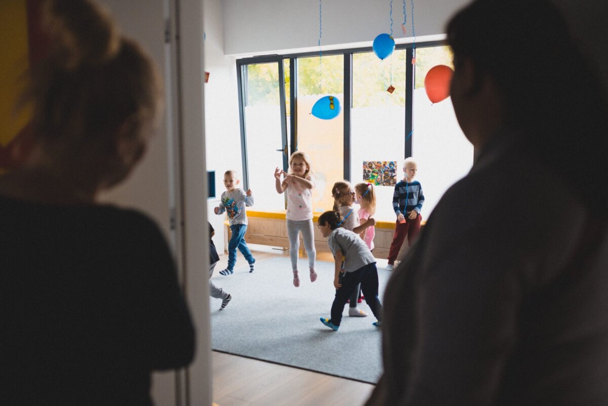 A group of children are playing and jumping around in a brightly lit room with large windows. Two adults stand in the doorway watching them. The room, ready for any event photography, is decorated with hanging balloons and other festive items.  