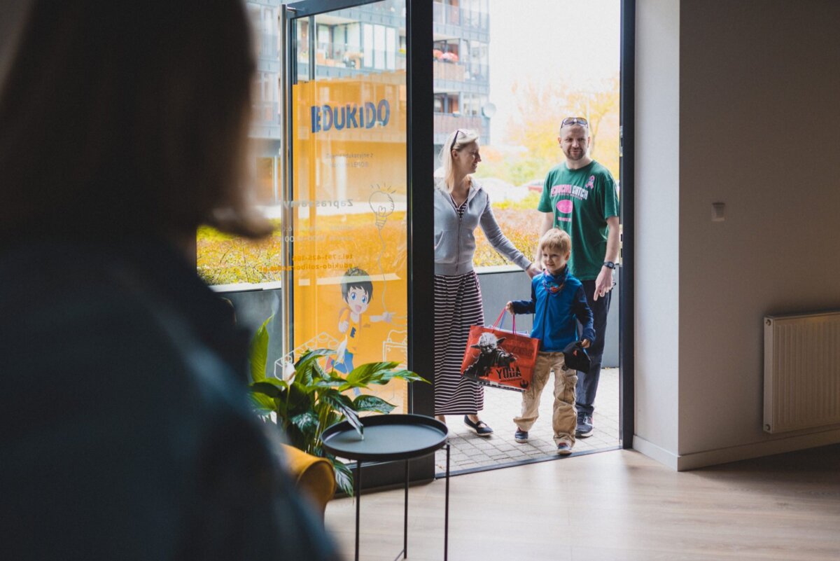 A family consisting of an adult man, an adult woman and a young child with a toy truck enters a bright room with large windows. In the foreground, another person is partially visible greeting them. Modern furniture and plants complete the scene, perfect for any photographic event.  