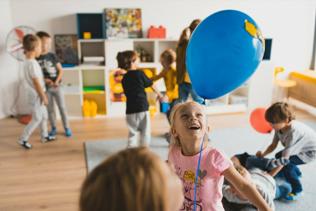 A group of children play in a room with a wooden floor and white shelves filled with toys. A girl in a pink shirt smiles and holds a blue balloon, beautifully captured by event photographer Warsaw. Other children interact and play with the toys in the background.  