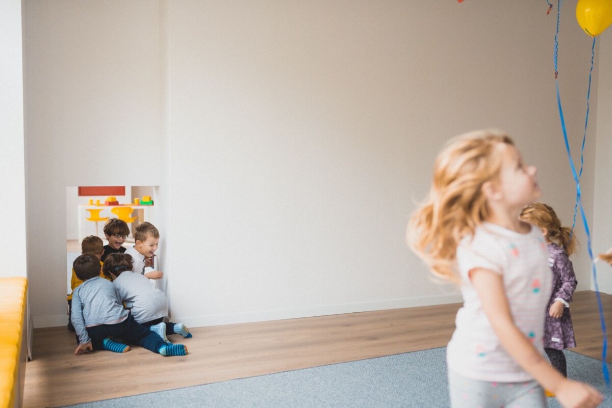 A group of young children sit on the floor near a small playground in the corner of the room. One child walks by in the foreground. The walls are white, and the floor is a mix of light wood and blue carpet. Balloons and streamers decorate the space, perfect for an event photography session capturing joyful moments.   