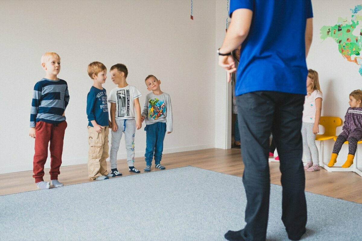 A group of young children are standing in a classroom listening to an adult in a blue shirt and black pants. Five children stand in a row to the left, while three others sit on a bench to the right. In a bright, colorful room, a map of the world hangs on the wall, perfectly captured by event photographer Warsaw.  