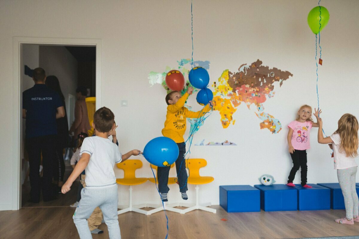 Children play with colorful balloons in a room decorated with a world map mural, creating a perfect photo-op of the events. Some children are standing, while others are sitting on blue and yellow chairs. In the doorway stands a person wearing a T-shirt that says "INSTRUCTOR." The atmosphere is lively and fun.   