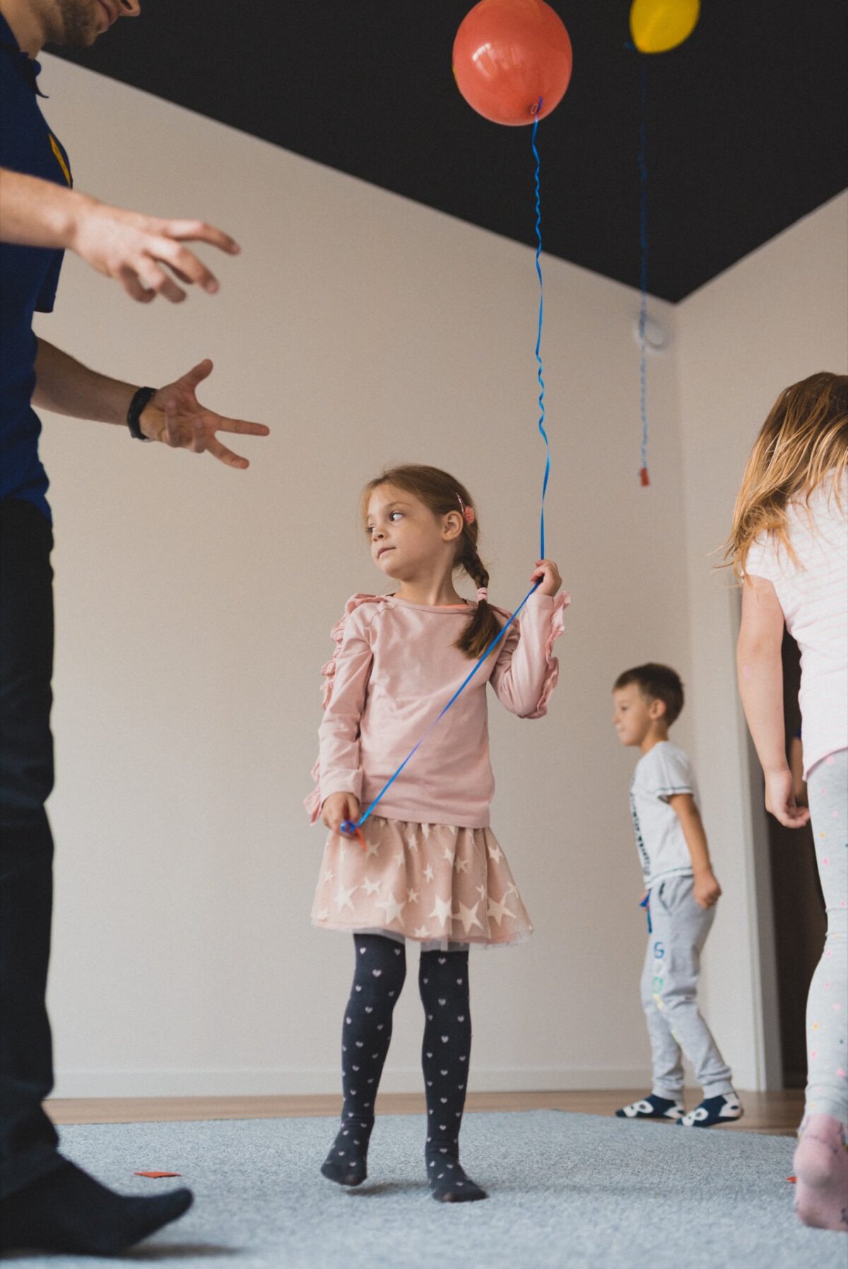 A young girl in a pink skirt and black tights holds a balloon on a string, looking up. An adult stands nearby with his arms outstretched, while another child plays in the background. The location of the action is an interior with a simple white wall, perfect for photographing events and capturing every moment.  