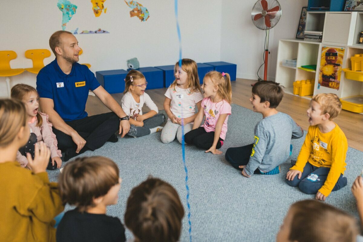 The teacher sits on the floor in a circle with young children in the classroom. The children seem engaged and are smiling as they participate in the blue string activity. The room has colorful chairs, educational maps on the wall and various storage containers, beautifully captured by event photographer Warsaw.  