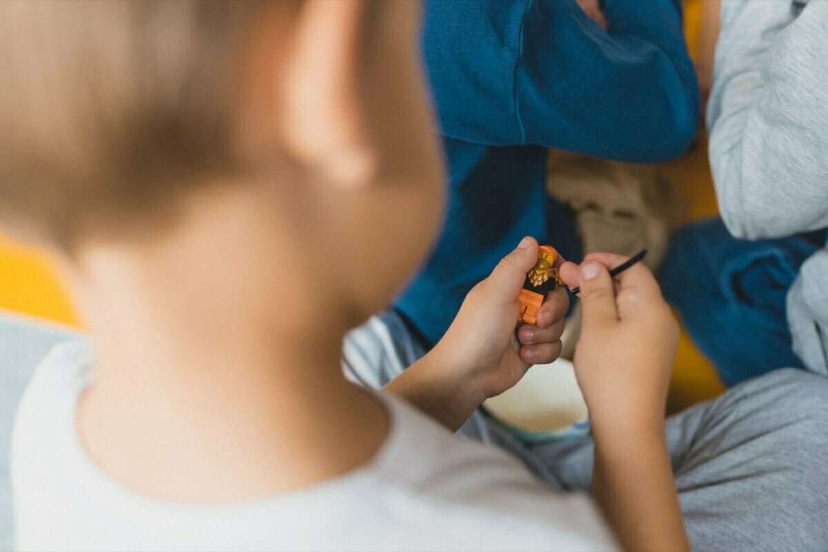 From behind, a child can be seen holding and watching a small toy figure. The child is wearing a white shirt, around him you can partially see people dressed in blue-gray clothes. The background is out of focus, the focus is on the child and the toy - in true professional event photography style.  