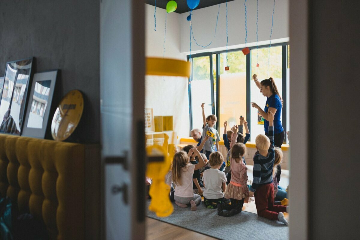 A group of young children sit on the floor in a circle around an adult instructor in a bright, colorful room. Captured by *Warsaw event photography*, the instructor raises an object, and the children eagerly raise their hands. The space is decorated with balloons and streamers.  