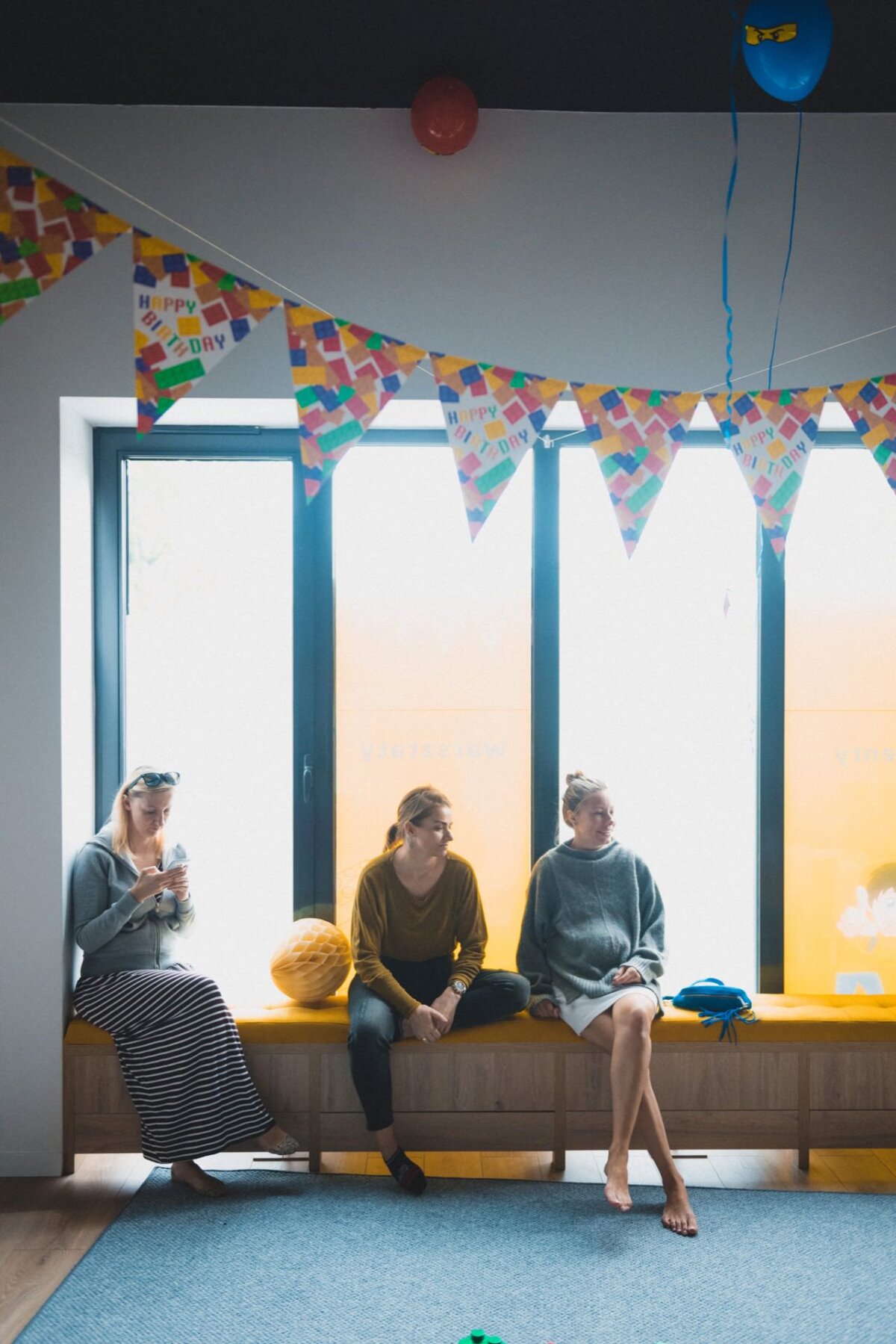 Three people are sitting on a yellow bench in a room decorated with colorful triangular birthday banners. One person is looking at his phone, another is sitting with his arms folded, and another is barefoot. Large windows behind them let in natural light, perfect for event photography.  