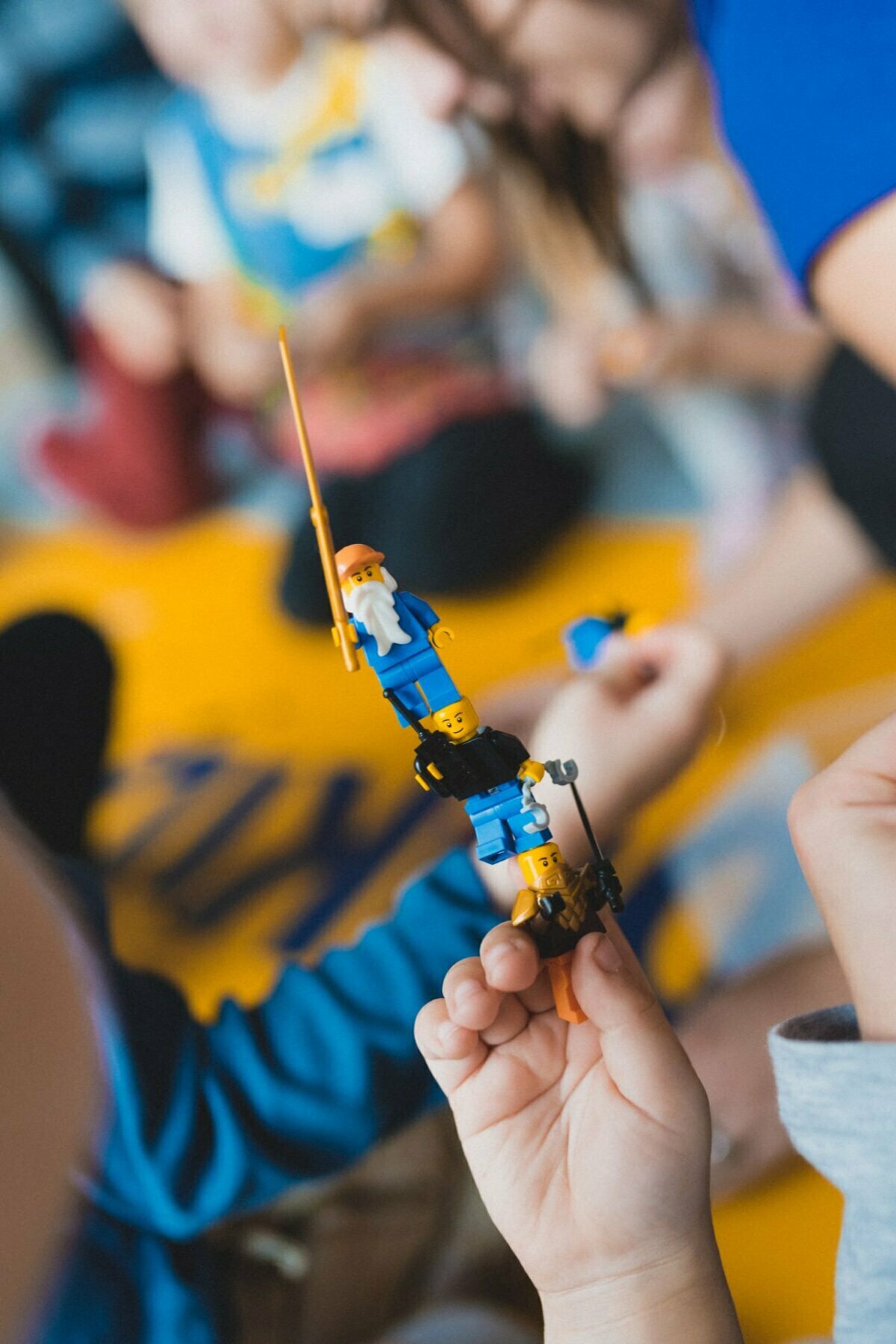 The child is holding a stack of LEGO minifigures, one on top of the other. The characters are dressed in blue and yellow, one with a white beard and a walking stick. Against a blurry background, other children are playing among the yellow and blue elements. This scene perfectly captures the essence of event photography.   