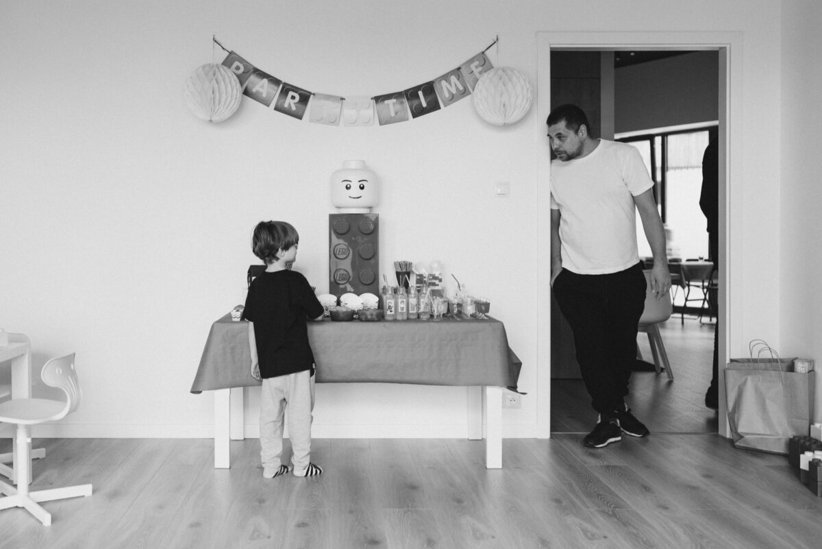 A child stands in front of a table decorated for the event with a LEGO theme, including a large LEGO figure. A banner with the words "PART-TIME WORK" hangs above the table. An adult looks at a child from a nearby walkway. Captured in black and white, this event photography perfectly captures the joyous moment.   