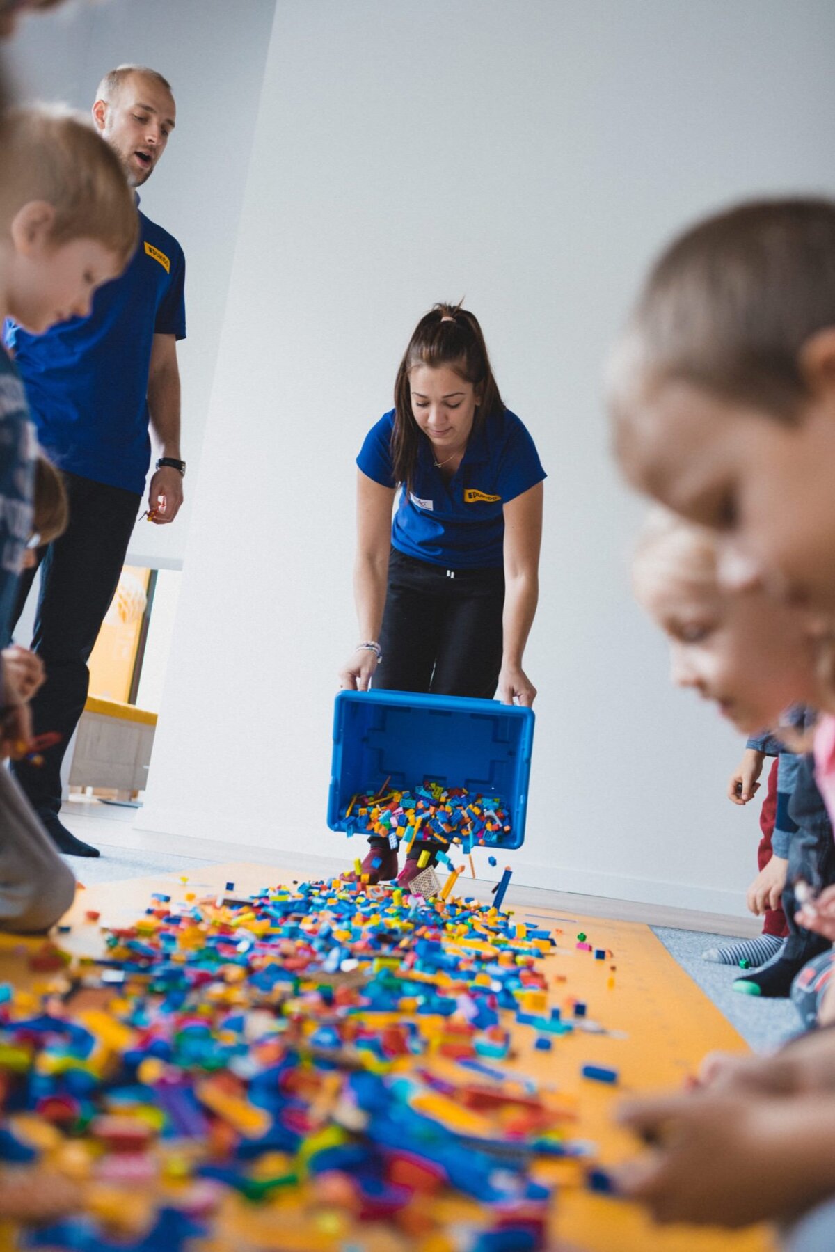 A person wearing a blue shirt pours a large container of colorful LEGO bricks onto the floor, while several children gather around, reaching for the bricks. Another person, also wearing a blue shirt, stands in the background. The scene is perfectly captured, perfect for event photography in a bright room.  